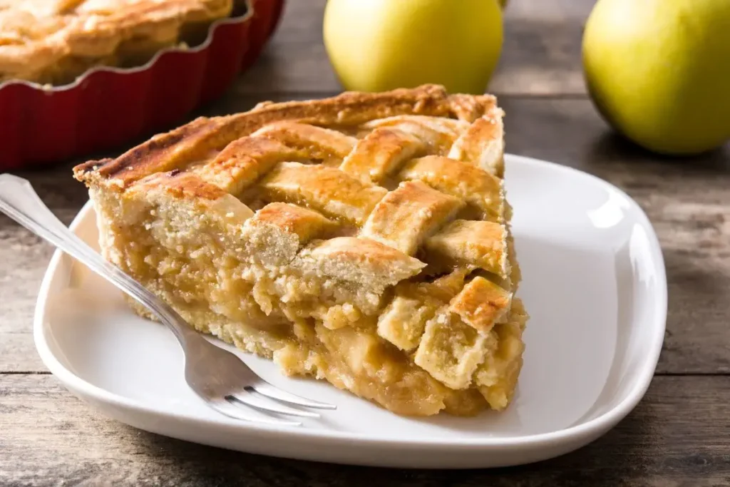 A slice of apple pie on a white plate with green apples in the background.