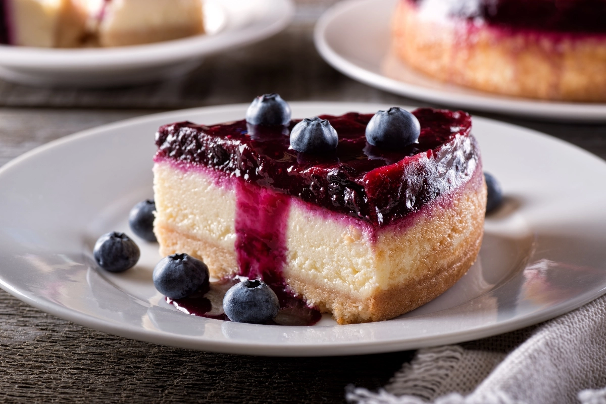 A slice of blueberry cheesecake cake on a white plate, topped with fresh blueberries and a glossy blueberry sauce.