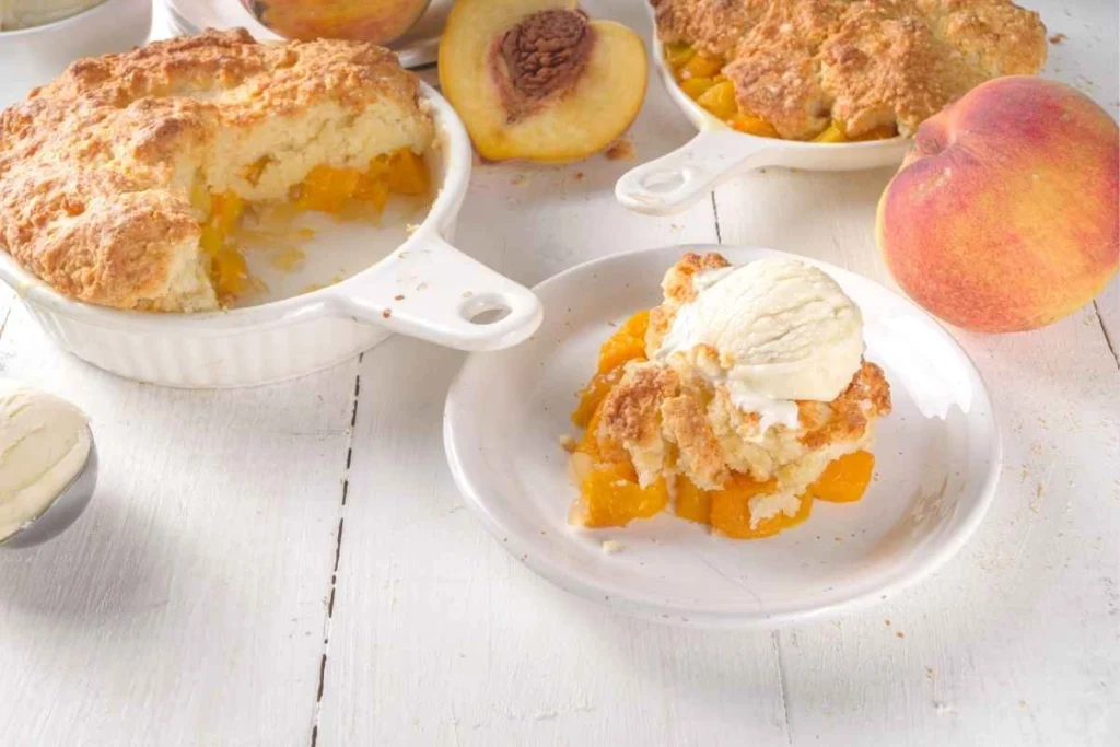 Freshly baked peach cobbler served in a white dish, with a portion plated and topped with a scoop of vanilla ice cream.