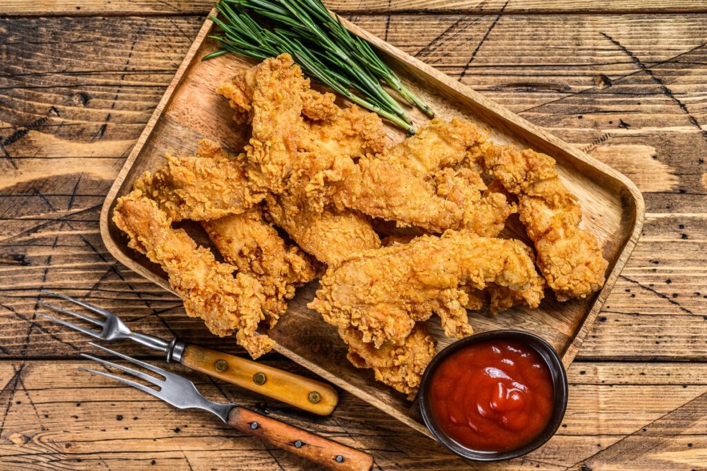 Crispy golden fried chicken tenders served on a wooden tray with ketchup and fresh herbs.