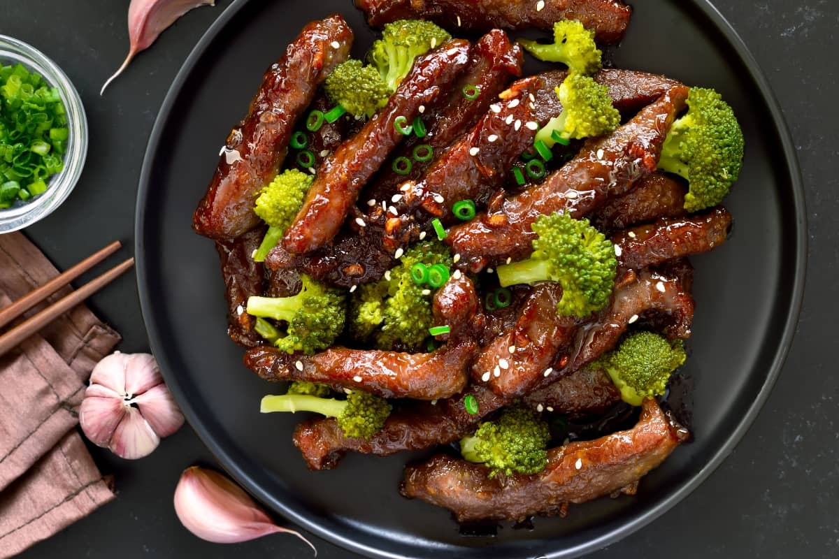 Chinese beef and broccoli stir-fry with sesame seeds and green onions on a black plate.