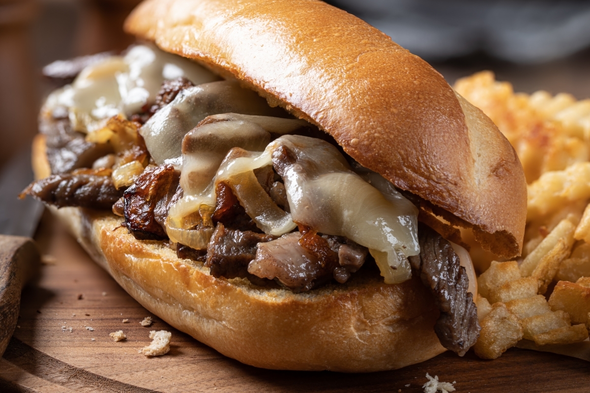 A Philly cheesesteak sandwich with melted cheese, sautéed onions, and tender beef on a toasted roll, served with waffle fries.