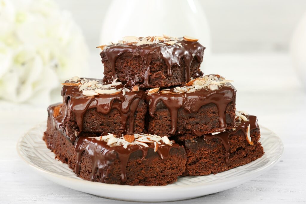 Rich chocolate brownies with a glossy chocolate glaze, topped with almond flakes, stacked on a white plate.